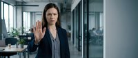 Serious woman with long dark hair, Caucasian ethnicity, wearing dark blazer and gray shirt, making stop gesture with raised hand in modern office corridor with glass partitions and natural light.