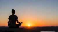 Woman silhouette meditating in lotus pose during golden sunset over mountains and lake creating peaceful zen atmosphere for wellness and mindfulness practice.