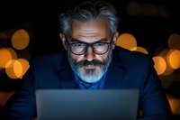 Mature businessman with gray beard and glasses working late on laptop with dramatic blue light illumination and bokeh background effect.