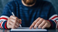 Young man in blue sweater with striped sleeves writing on paper with pen at desk for business planning and note-taking tasks.