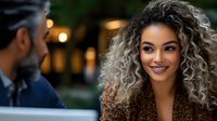 Young mixed race woman with curly blonde ombre hair smiling warmly during romantic evening date outdoors. Natural lighting creates soft bokeh background effect.