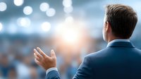 Professional businessman presenting to audience with bright stage lights and bokeh background for corporate presentations and public speaking events.