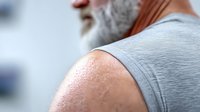 Senior Caucasian man with gray beard wearing sleeveless shirt, close-up view of muscular shoulder and neck showing skin texture and age details.