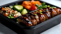 Glazed teriyaki chicken bento box with steamed rice, fresh cucumber, carrots, and garnished with sesame seeds and green onions served in black container.