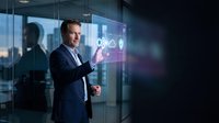 Professional businessman in dark suit presenting digital interface with holographic icons in modern glass office environment for corporate technology presentations.