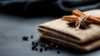 Aromatic cinnamon sticks and star anise on burlap cloth with scattered black peppercorns against dark background. Perfect for culinary themes.