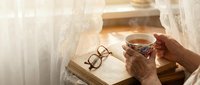 Cozy morning reading scene with hands holding steaming coffee cup, open book and eyeglasses on wooden surface near bright window with soft natural light.