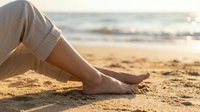 Woman relaxing barefoot on sandy beach at sunset with ocean waves in background, peaceful vacation moment for wellness and travel concepts.