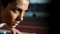 Determined female athlete with sweat droplets on face showing intense focus and concentration during training session on running track.