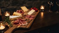 Gourmet charcuterie board with assorted meats, cheeses and nuts on wooden platter surrounded by flickering candles creating warm romantic dining atmosphere.
