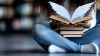 Young woman reading open book with white angel wings emerging from pages while sitting cross-legged in jeans, symbolizing knowledge, learning and imagination.