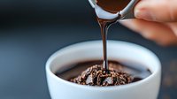 Liquid chocolate sauce pouring from spoon into white ceramic bowl, creating glossy stream and ripples on dark chocolate surface. Food photography macro shot.
