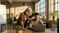 Mature woman with gray hair and younger man collaborating at laptop in bright office workspace with plants and natural light from industrial windows.