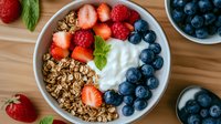 Fresh berries and granola breakfast bowl with yogurt, strawberries, blueberries, raspberries and mint leaves arranged on wooden table, healthy morning meal.
