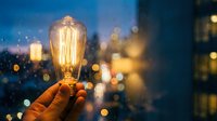 Hand holding glowing vintage Edison bulb against blurred city lights at dusk, symbolizing innovation, creativity and bright ideas for business concepts.