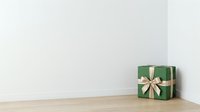 Green gift box with cream ribbon bow on wooden floor against white wall.