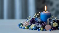 Blue candle burning surrounded by colorful gemstones crystals and decorative beads on white surface creating peaceful meditation atmosphere.