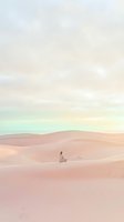 Woman in light clothing sitting alone on rolling pink sand dunes under soft pastel sky, evoking solitude, meditation, and desert landscape exploration for travel and wellness content.