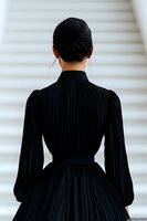 Back view of Asian woman in elegant black pleated dress with high collar and gathered waist against white background, hair styled in sleek updo with silver hoop earrings.