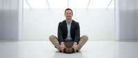 Professional businessman sitting cross-legged on white floor in modern minimalist office space, wearing dark blazer and khaki pants for corporate wellness.