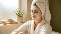 Young woman enjoying spa treatment with clay face mask and towel wrapped hair in bright bathroom for skincare routine and wellness relaxation.