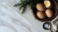 Fresh farm eggs in rustic woven basket with butter, sea salt and rosemary sprig on white marble countertop, overhead view of minimal breakfast composition.