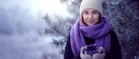 Young Caucasian woman in purple knit scarf and white beanie holding snow in cupped hands during winter day outdoors among frost-covered evergreen branches.