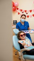 Young child wearing red sunglasses giving thumbs up during dental visit with female dentist in blue scrubs and mask. Heart decorations create festive atmosphere.