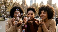 Three young African American women laughing and blowing soap bubbles in urban park with city skyline background during golden hour sunset.