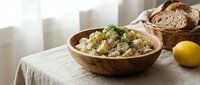 Wooden bowl filled with creamy potato salad garnished with fresh herbs, served with sliced bread and lemon on linen tablecloth for food styling and culinary content.