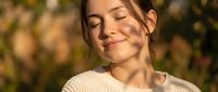 Young woman with long brown hair and fair complexion, wearing a cream-colored knit sweater, smiling with eyes closed in warm sunlight outdoors. Suitable for lifestyle, wellness, and happiness concepts.