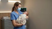 Female healthcare worker in blue scrubs and medical mask holding newborn baby in hospital nursery setting for medical and childcare concepts.