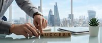 Professional businessman arranging wooden letter blocks spelling SUCCESS on modern office desk with city skyline view through window.
