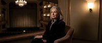 Elegant mature businesswoman with blonde hair sitting confidently in luxurious hotel lobby or upscale restaurant interior with warm ambient lighting.