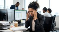 Stressed African American businesswoman experiencing headache while working at computer in busy corporate office environment with colleagues in background.