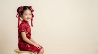 Young Asian girl, approximately 5-7 years old, wearing traditional red silk outfit with floral hair accessories and ribbons, sitting pose against neutral background for cultural celebrations.
