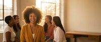 Confident African American businesswoman with curly hair smiling in modern office meeting room with diverse team of female colleagues in background.