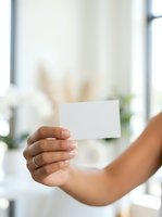 Close-up of woman's hand holding blank white business card in bright office environment with blurred background, suitable for corporate branding and marketing mockups.