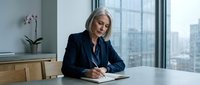 Mature woman with gray hair, Caucasian ethnicity, wearing dark business blazer and blue shirt, writing in notebook at modern office desk with city skyline view through floor-to-ceiling windows, professional workplace setting.