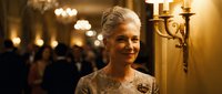 Elegant senior woman with silver hair smiling warmly at formal evening event with golden lighting and ornate decor in luxurious ballroom setting.