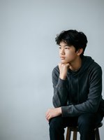 Young East Asian man in casual charcoal hoodie sitting thoughtfully on wooden stool against white background, hand to chin in contemplative pose for portrait and lifestyle use.