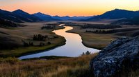 Meandering river reflects golden sunset light as it winds through mountain valley, with silhouetted peaks and rolling grasslands creating dramatic wilderness landscape.