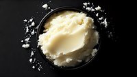 Fresh creamy mashed potatoes in black bowl with butter melting on top, scattered flour on dark surface for cooking and recipe preparation.