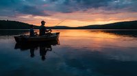 Silhouetted male angler fishing from small boat on calm lake at sunset, golden sky reflecting on water surface, peaceful outdoor recreation scene.