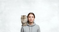 Young woman with brown hair and fair complexion wearing gray hoodie, posing with fluffy tabby cat on shoulder against white textured background. Studio portrait for pet, lifestyle, and animal companionship concepts.
