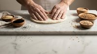 Hands kneading dough on floured surface with wooden bowls of seeds and grains for homemade bread baking in rustic kitchen setting.