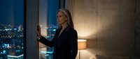 Elegant senior Caucasian businesswoman in dark suit standing by window in luxury hotel room with city lights at night, thoughtful expression.