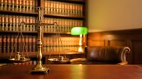 Golden scales of justice on wooden desk in law library with legal books and green banker lamp creating warm professional atmosphere for legal services.