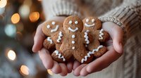 Smiling gingerbread cookies with white icing decorations held in cozy sweater-clad hands against blurred holiday lights background, creating festive mood.