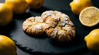Crinkled lemon cookies dusted with powdered sugar on dark slate with fresh lemons, perfect for bakery menus and dessert photography.